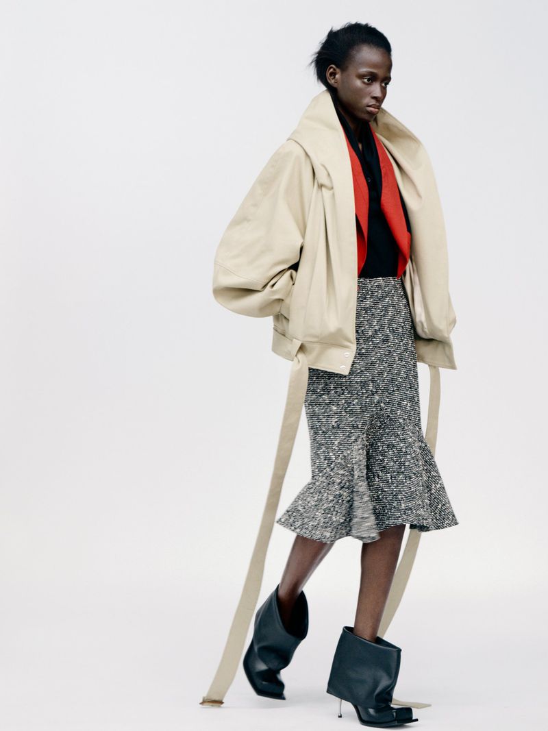 Elegant studio shot of a Black female fashion model in a beige jacket, patterned skirt, and black boots against a clean white backdrop.