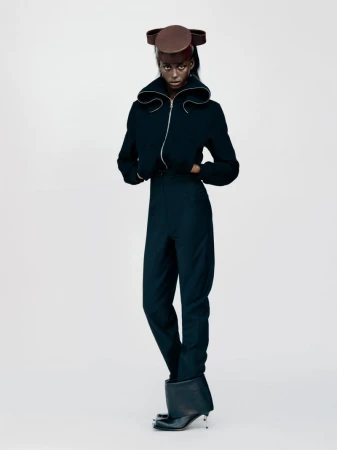 A Black model wearing a structured black zip-up jacket, trousers, and a sculptural brown hat against a plain white studio background.