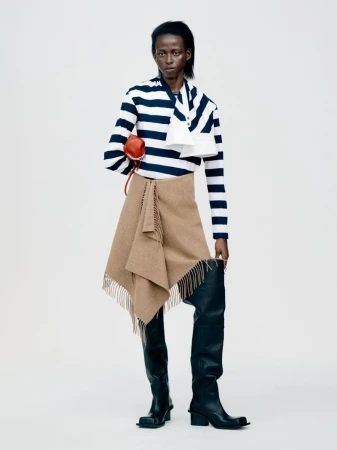 Model wearing striped top, tan fringed skirt, and tall black boots against a white studio background