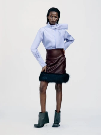 Model wearing a light blue bow-collar shirt, dark leather skirt with faux fur trim, and black boots against a white backdrop
