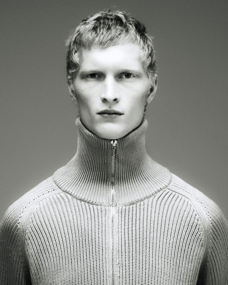 Close-up of a young adult male model with blonde hair wearing a zipped-up, high-neck knit sweater, posing in a studio with a neutral background.