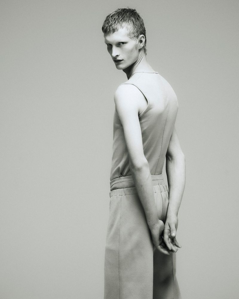 Monochrome fashion shot of a young adult male model wearing light-colored sleeveless top and cropped pants, turning his head to face the camera.