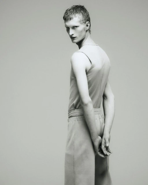 Monochrome fashion shot of a young adult male model wearing light-colored sleeveless top and cropped pants, turning his head to face the camera.