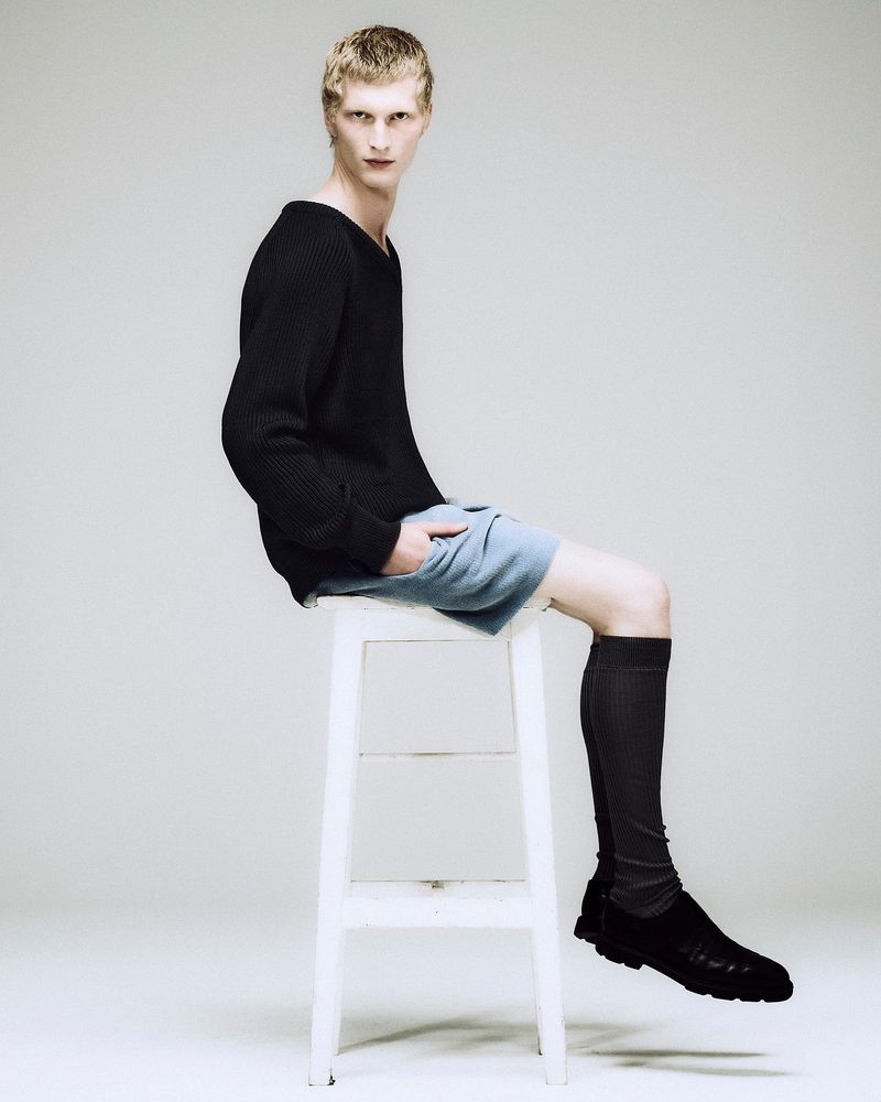 Male fashion model sits on a white stool in a studio, wearing a black sweater, blue shorts, black socks, and black shoes against a white backdrop.