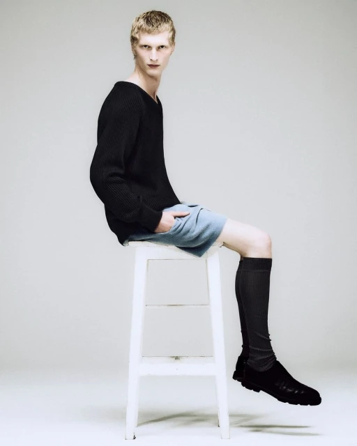 Male fashion model sits on a white stool in a studio, wearing a black sweater, blue shorts, black socks, and black shoes against a white backdrop.