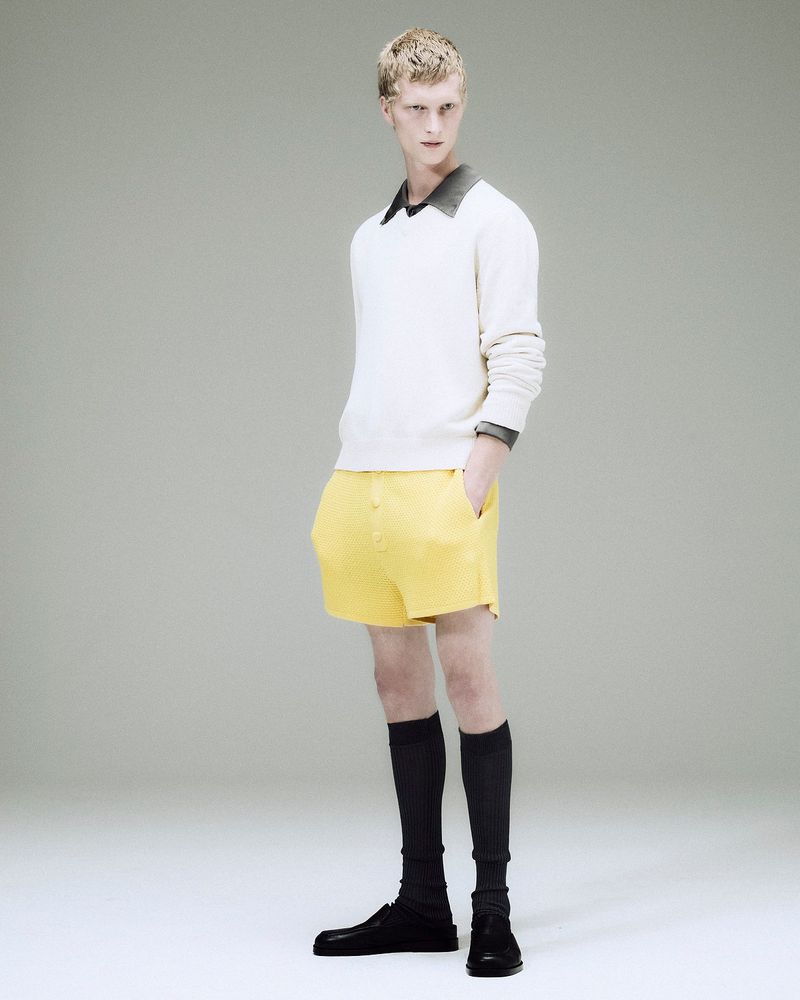 Model standing against a light grey seamless background wearing a white sweater, yellow shorts, and black knee-high socks.