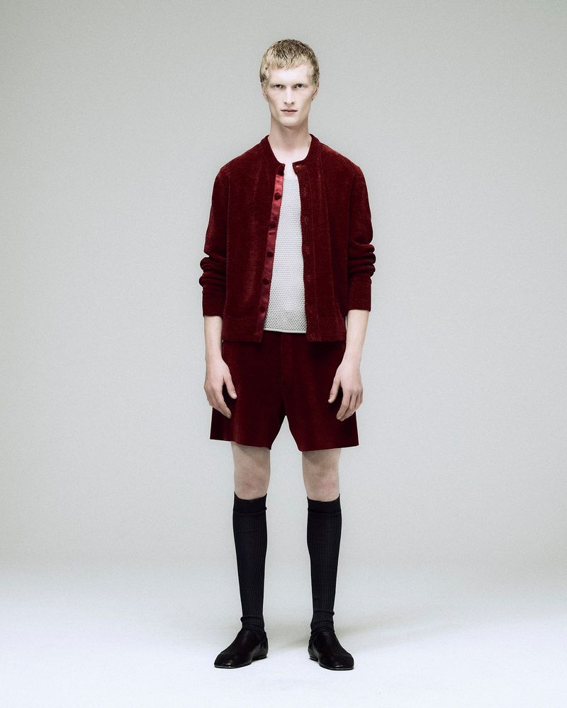 Male model in a maroon velvet jacket, shorts, black socks, and shoes poses against a white backdrop in a full-body studio shot.