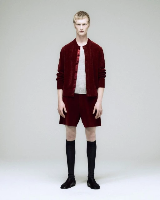 Male model in a maroon velvet jacket, shorts, black socks, and shoes poses against a white backdrop in a full-body studio shot.