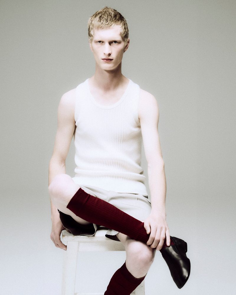 Male model seated on a white stool wearing a white tank top and dark knee-high socks against a neutral studio background