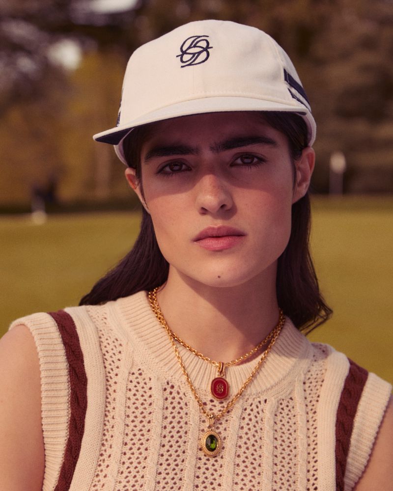 Close-up of a woman wearing a cap and vintage-inspired vest with two gold necklaces with red and green gemstones on a lawn.