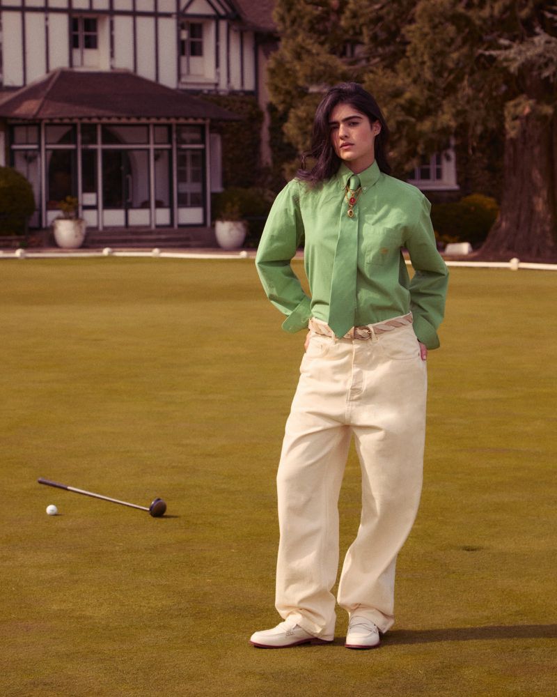 Woman in green shirt and cream pants poses with golf club and ball on a green lawn in front of a large house on a sunny day.