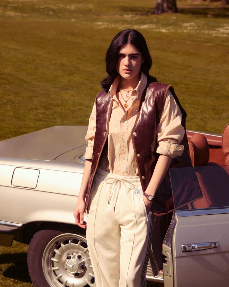 A woman in a vest stands near a vintage car on a sunny day, wearing casual attire and jewelry, in a grassy outdoor setting.