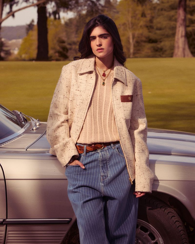 Brunette woman in a cream tweed jacket and blue striped pants leans against a silver car in a grassy outdoor setting on a sunny day.