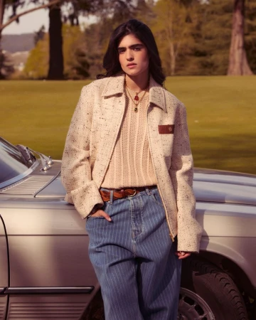Brunette woman in a cream tweed jacket and blue striped pants leans against a silver car in a grassy outdoor setting on a sunny day.