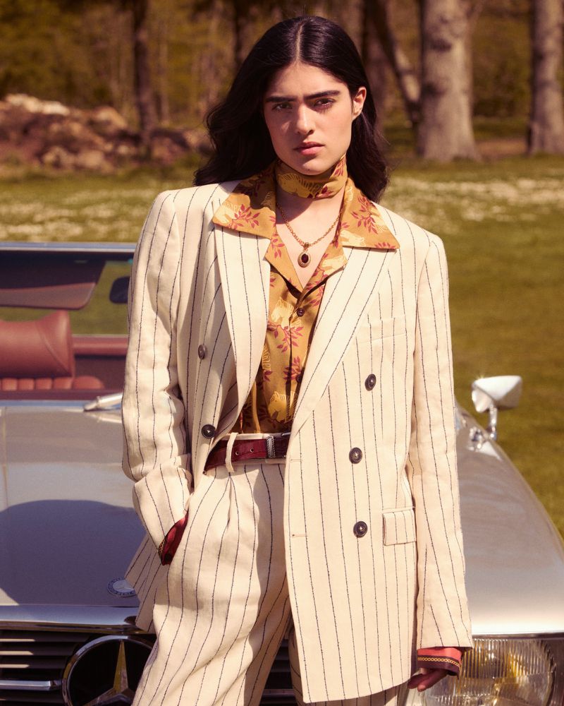 Woman in a striped suit poses with classic car in grassy, outdoor setting with trees in the background. Fashion editorial photography.