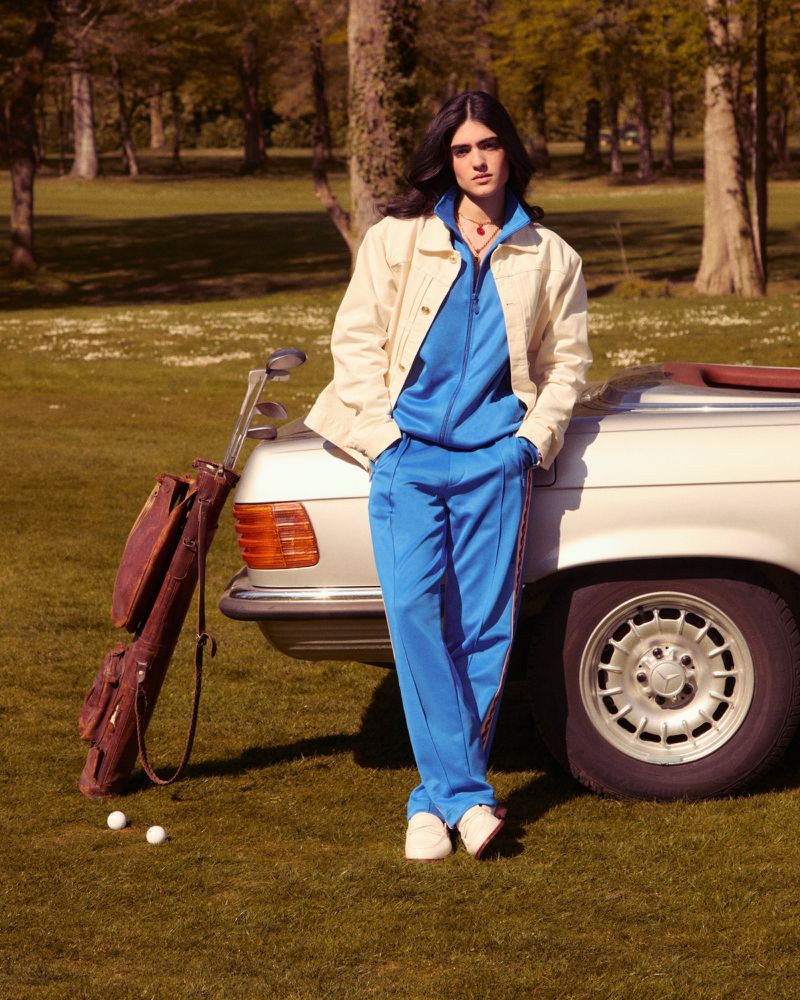 Young man in a blue tracksuit leaning on a vintage car, posing on a sunny lawn with golf balls and a golf club bag; outdoor fashion shoot.