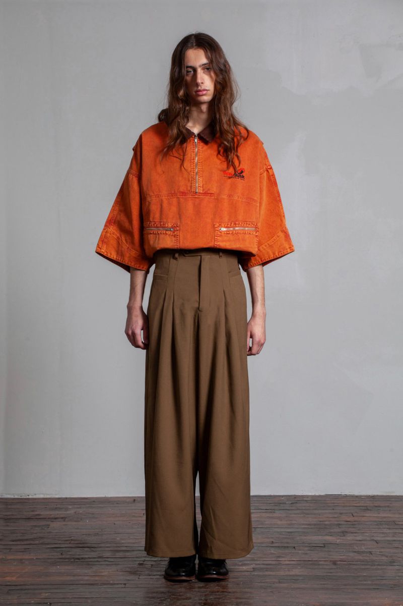 Studio shot of a slender adult male model with long brown hair, wearing a bright orange shirt and wide-legged khaki trousers.