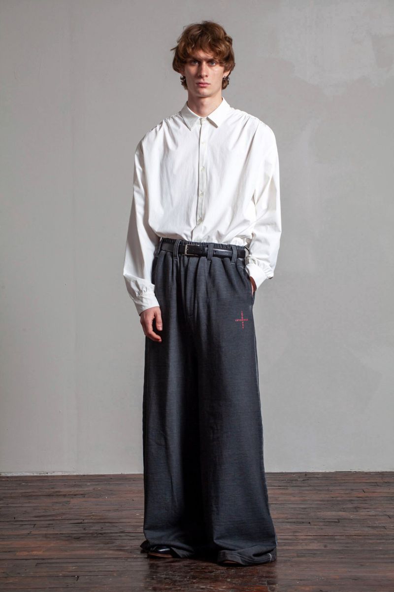 Model in white long-sleeve shirt and dark grey wide trousers standing against a neutral grey studio background