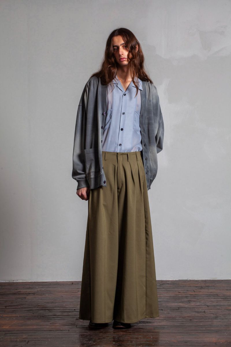 Full-length shot of a slender adult man in wide-leg olive trousers, light blue button-down shirt, and a faded grey cardigan, posing indoors.