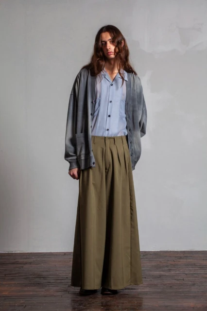 Full-length shot of a slender adult man in wide-leg olive trousers, light blue button-down shirt, and a faded grey cardigan, posing indoors.