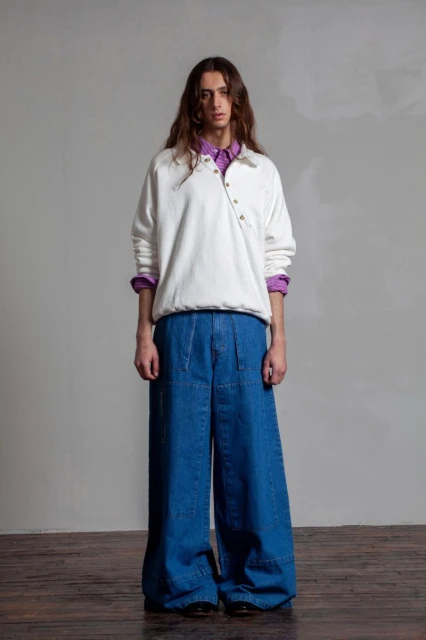 Full-body portrait of a young adult with long brown hair wearing a white sweatshirt, purple shirt, and oversized blue jeans in a studio.