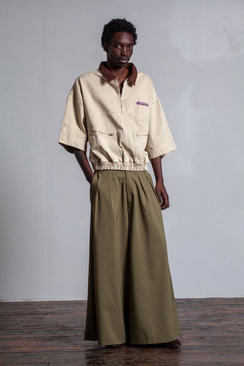 An adult Black male model poses in an earth-toned tan polo shirt and large olive-green trousers in a full-length indoor fashion shot.