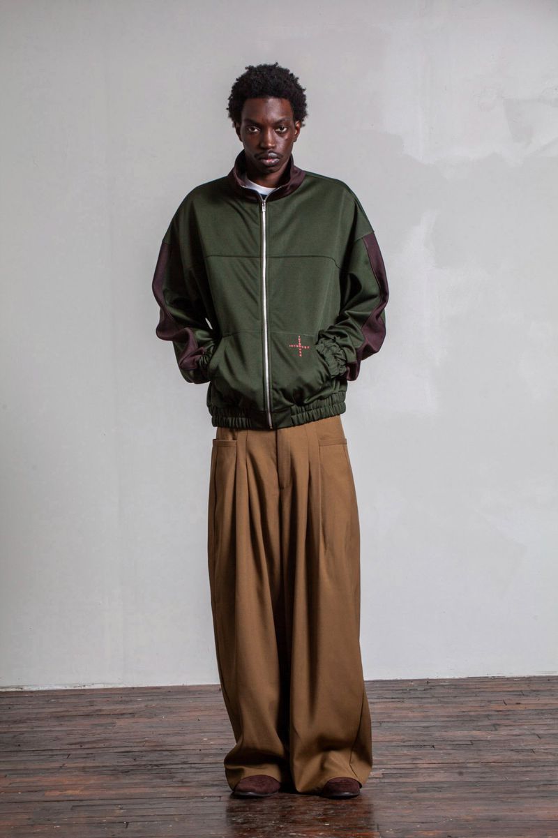 Model wearing a green zip jacket and brown pleated trousers standing against a pale wall and dark wooden floor.