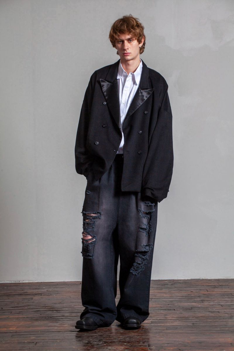 Adult male fashion model wears oversized black blazer, white shirt, and distressed wide-leg jeans in a full-length studio shot.
