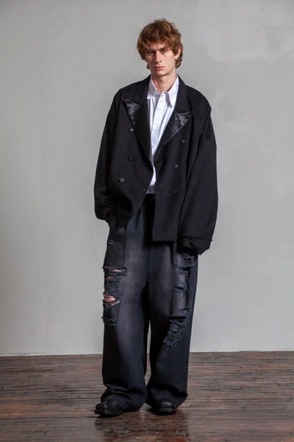 Adult male fashion model wears oversized black blazer, white shirt, and distressed wide-leg jeans in a full-length studio shot.