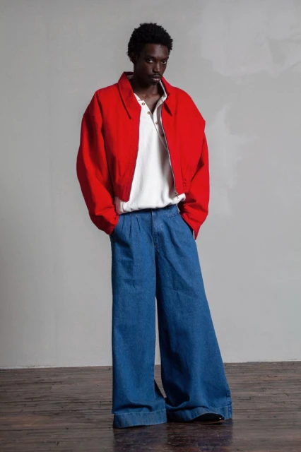 A stylish Black male model poses in a studio wearing a red jacket, white polo shirt, and wide-legged blue denim jeans.