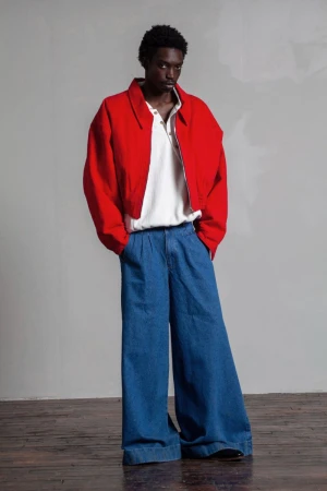 A stylish Black male model poses in a studio wearing a red jacket, white polo shirt, and wide-legged blue denim jeans.