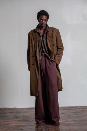 Model wearing a brown coat and dark trousers standing against a textured wall and wooden floor background.