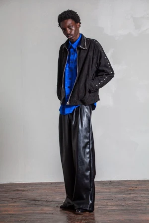 A man wearing a black studded jacket over a blue grid-patterned shirt and wide-leg black trousers standing against a plain white wall.