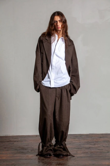 A person with long brown hair poses in a brown suit and white collared shirt against a grey studio backdrop, standing on a wooden floor.
