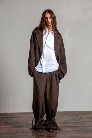 A person with long brown hair poses in a brown suit and white collared shirt against a grey studio backdrop, standing on a wooden floor.