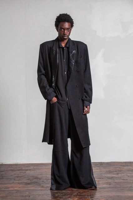 A Black male fashion model poses in a black, oversized coat and wide-leg trousers against a mottled grey background, studio lit.