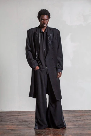 A Black male fashion model poses in a black, oversized coat and wide-leg trousers against a mottled grey background, studio lit.