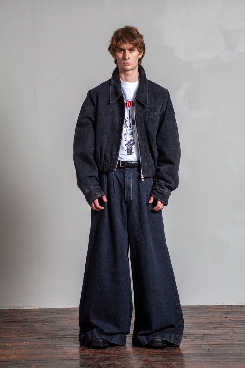 Man in wide-leg denim pants and a matching bomber jacket stands indoors against a textured gray wall and wooden floor.
