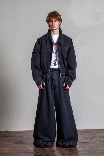 Man in wide-leg denim pants and a matching bomber jacket stands indoors against a textured gray wall and wooden floor.