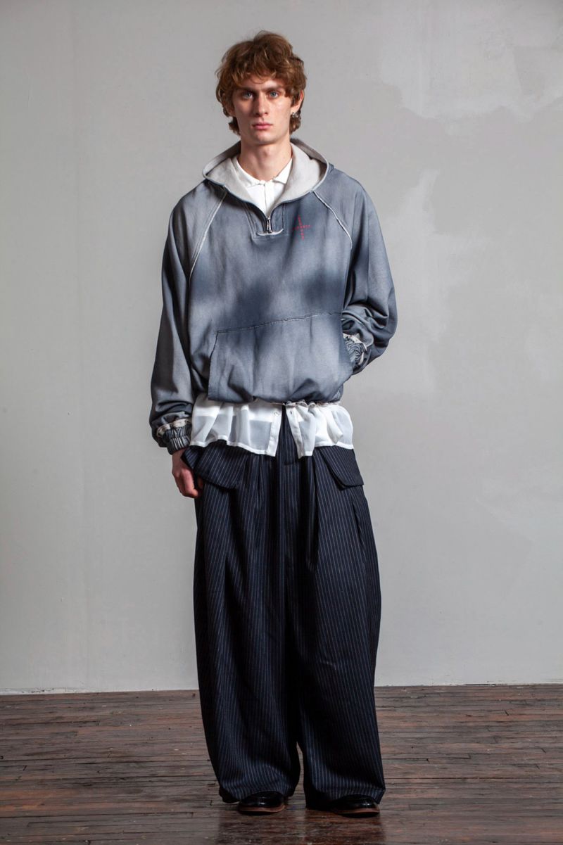 Full-length shot of an adult male fashion model wearing an oversized hoodie and wide-legged striped pants in a photo studio setting.