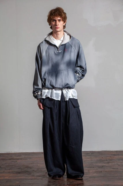 Full-length shot of an adult male fashion model wearing an oversized hoodie and wide-legged striped pants in a photo studio setting.
