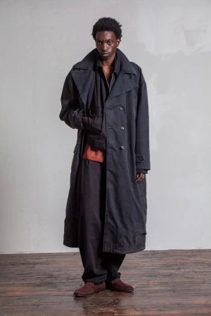 A man wearing an oversized charcoal grey trench coat over dark layers standing against a minimalist light grey wall on a wooden floor.