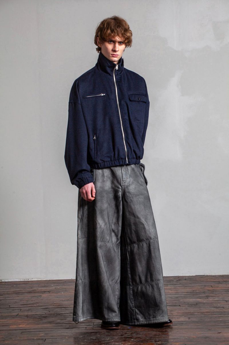 Model wearing a navy blue zip-up jacket and wide grey trousers standing against a pale grey studio wall.