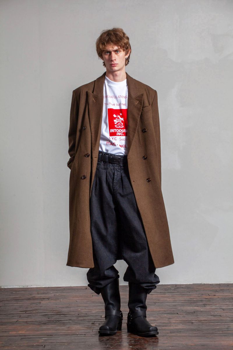 Adult man posing in a brown overcoat, t-shirt with graphic, black pants, and black boots against a neutral backdrop in a studio setting.