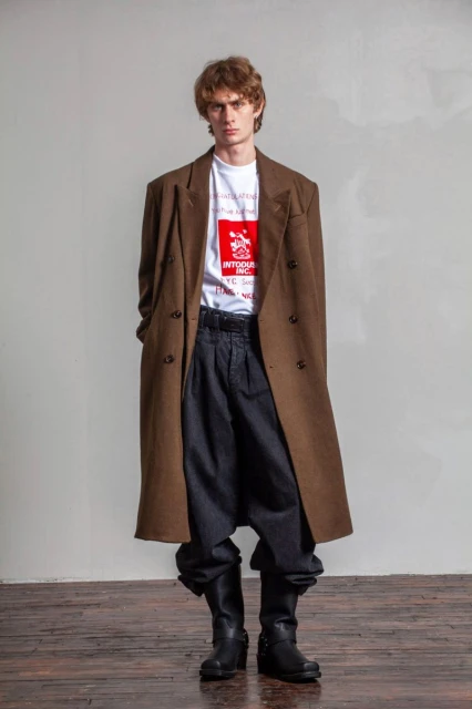 Adult man posing in a brown overcoat, t-shirt with graphic, black pants, and black boots against a neutral backdrop in a studio setting.