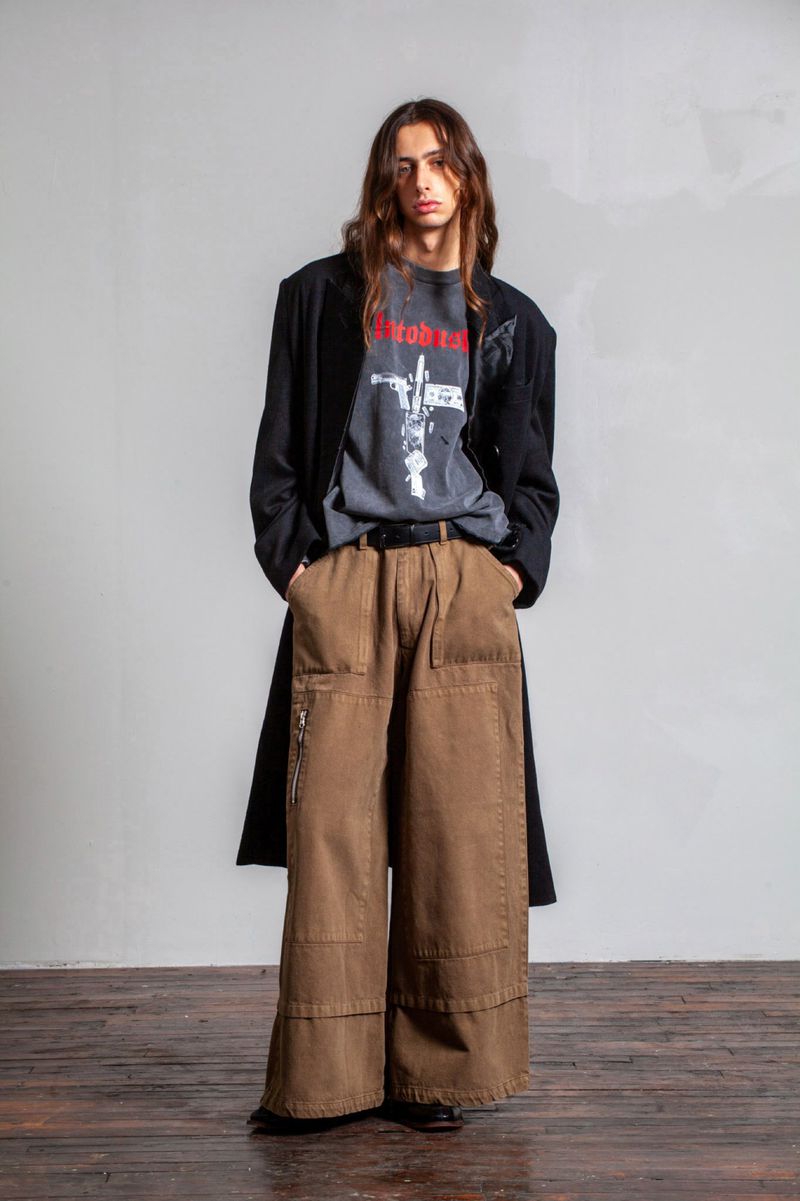 Full-length shot of a male model with long brown hair wearing a graphic tee, brown wide-legged pants, and a black coat.