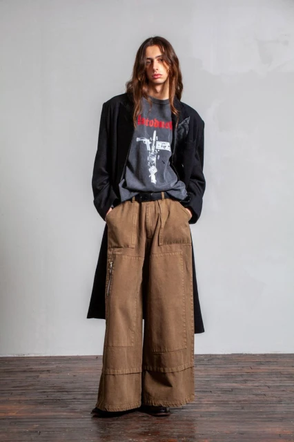 Full-length shot of a male model with long brown hair wearing a graphic tee, brown wide-legged pants, and a black coat.