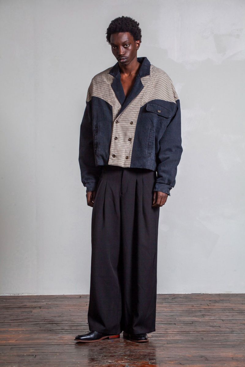 Dark-skinned male model in a double-breasted jacket and oversized black pants poses in a studio with a neutral grey background.