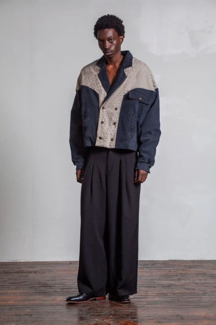 Dark-skinned male model in a double-breasted jacket and oversized black pants poses in a studio with a neutral grey background.