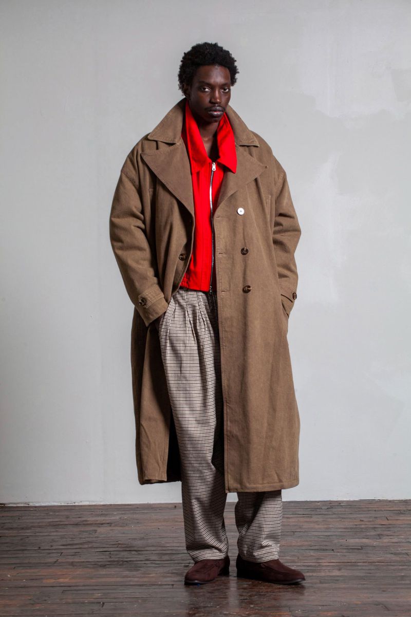 Man in a tan trench coat and patterned pants standing indoors against a grey wall and a dark wood floor. Fashion editorial.